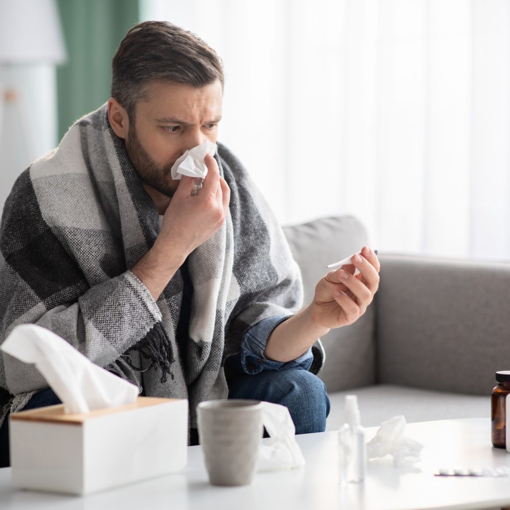 Man covered in blanket sneezing nose and checking for fever